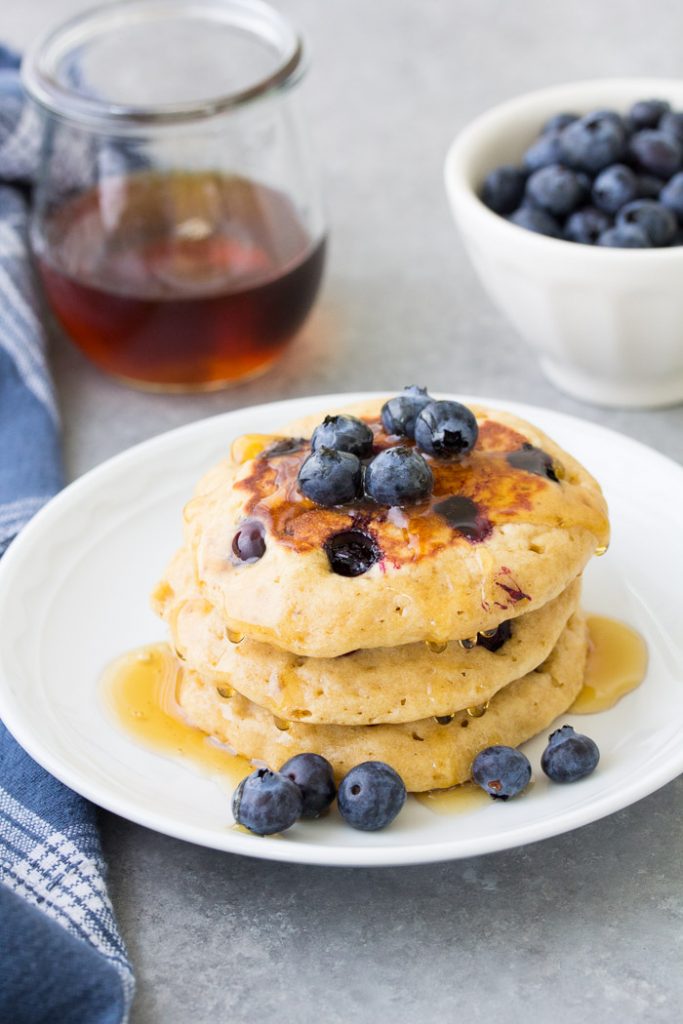 This easy blueberry pancakes recipe makes the best fluffy blueberry pancakes from scratch! These homemade pancakes are healthy, with just the right amount of sweet, juicy blueberries in them. Use fresh or frozen blueberries. They are perfect for a weekend breakfast! A stack of homemade blueberry pancakes on a plate with maple syrup and blueberries.