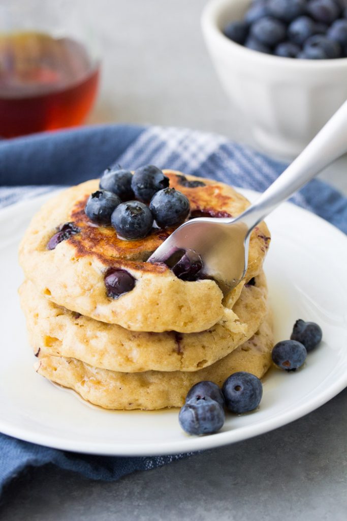 This easy blueberry pancakes recipe makes the best fluffy blueberry pancakes from scratch! These homemade pancakes are healthy, with just the right amount of sweet, juicy blueberries in them. Use fresh or frozen blueberries. They are perfect for a weekend breakfast! Taking a bite of blueberry pancakes with a fork.