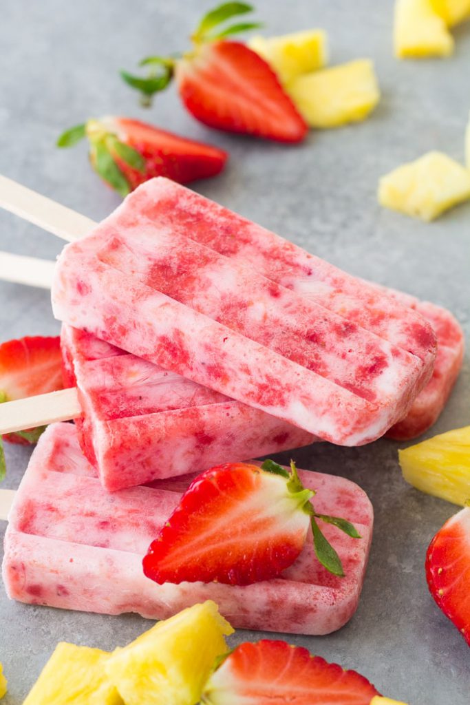 These Fruit and Yogurt Popsicles are easy to make and bursting with fresh fruit flavor! This homemade strawberry popsicle recipe is made with only four ingredients. These healthy popsicles are a fun summer treat for kids! A stack of homemade fruit and yogurt popsicles with strawberries and pineapple.