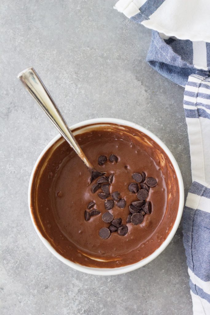 An easy chocolate mug cake that tastes like a brownie in a cup. This chocolate cake in a mug is a delicious microwave dessert that even a kid can make! This recipe has no egg and cooks in just 1 minute! Batter for a chocolate mug cake, in a white bowl with chocolate chips and a spoon.